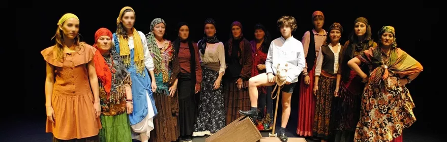 Grupo de actores con vestuario colorido posando en fila sobre un escenario oscuro.