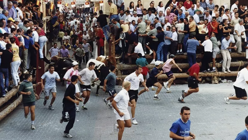 Multitud observa y corredores huyen de un toro por calle estrecha.