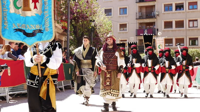 Desfile festivo con trajes tradicionales y estandartes en una calle urbana.