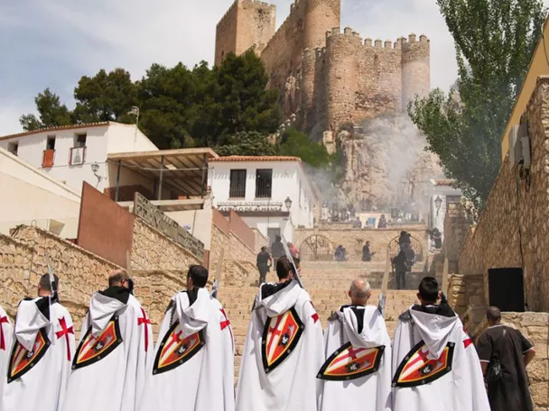 Grupo de personas con capas blancas avanzando hacia una fortaleza entre humo.