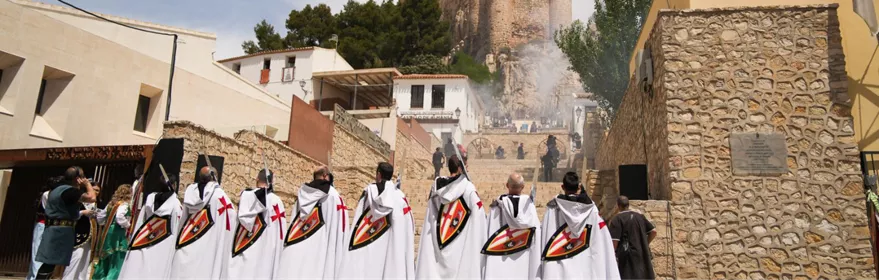 Grupo de personas con capas blancas avanzando hacia una fortaleza entre humo.