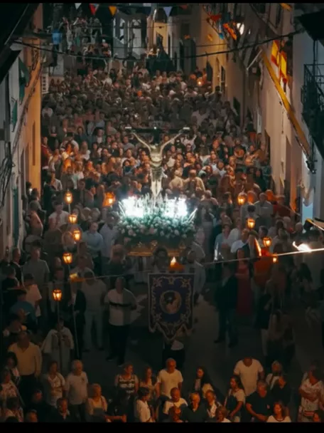Procesión nocturna con multitud portando luces por una calle estrecha.