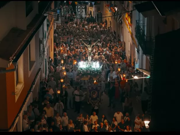 Procesión nocturna con multitud portando luces por una calle estrecha.