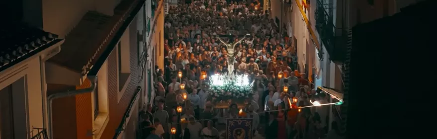 Procesión nocturna con multitud portando luces por una calle estrecha.