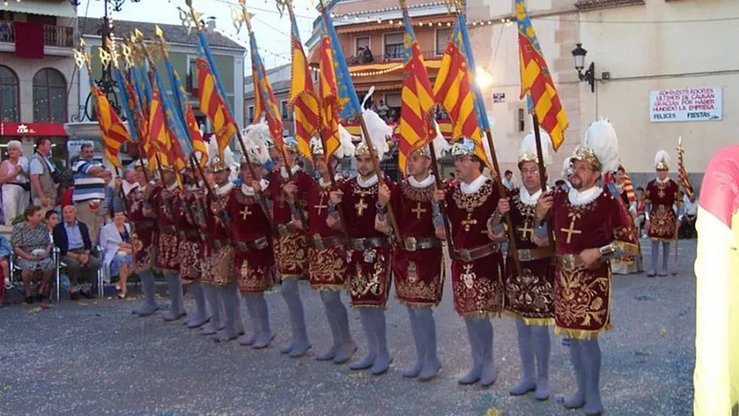 Fila de personas vestidas con trajes históricos portando banderas en una plaza.