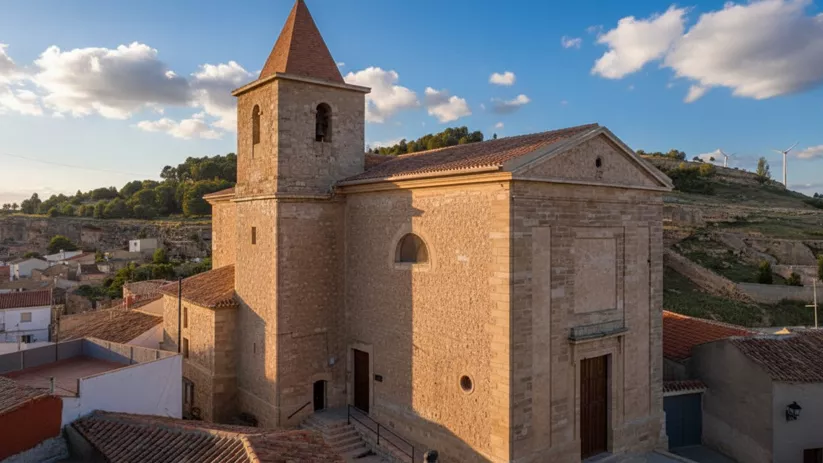 Iglesia de piedra en Higueruela con torre campanario, situada entre casas tradicionales y colinas al fondo.