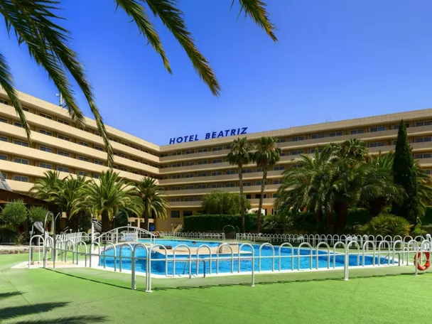 Fachada de hotel moderno con jardín de palmeras y piscina exterior.