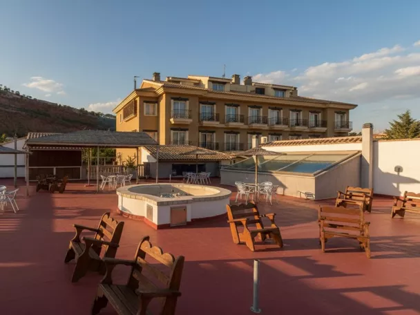 Terraza amplia con mesas y bancos, con edificio de varias plantas y colinas al fondo.