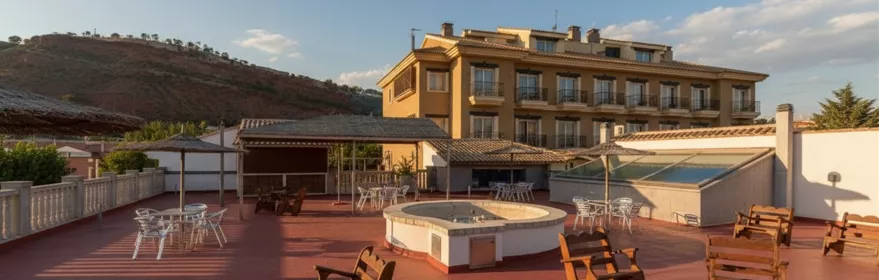 Terraza amplia con mesas y bancos, con edificio de varias plantas y colinas al fondo.