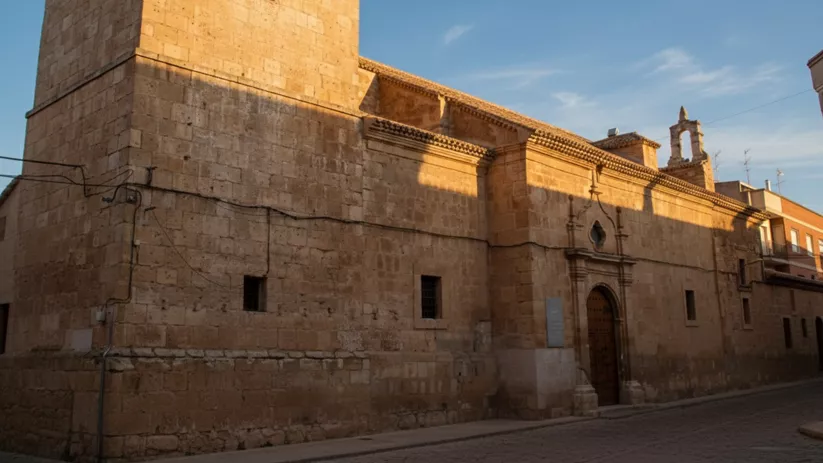 Fachada lateral de iglesia de piedra con torre cuadrada y portada, iluminada por el sol.