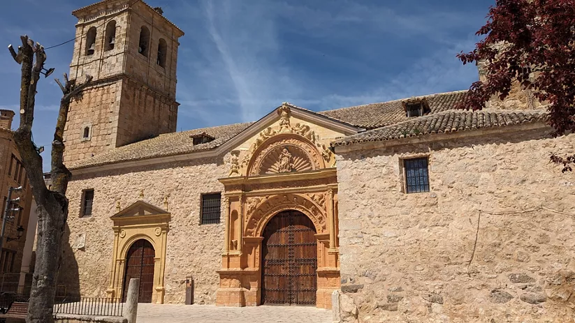 Vista frontal de iglesia de piedra con torre cuadrada y portada decorada en plaza abierta.