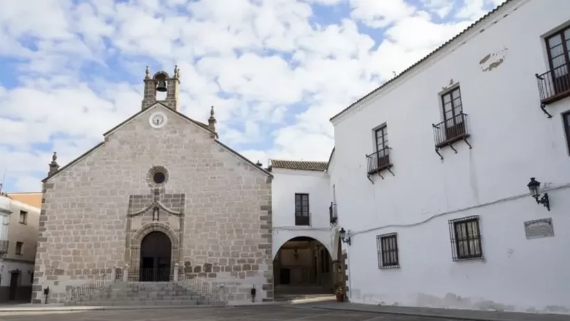 Iglesia de piedra frente a una plaza vacía junto a edificios blancos.