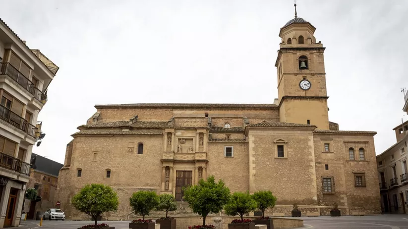 Fachada de iglesia de piedra con torre y reloj en una plaza con jardineras.