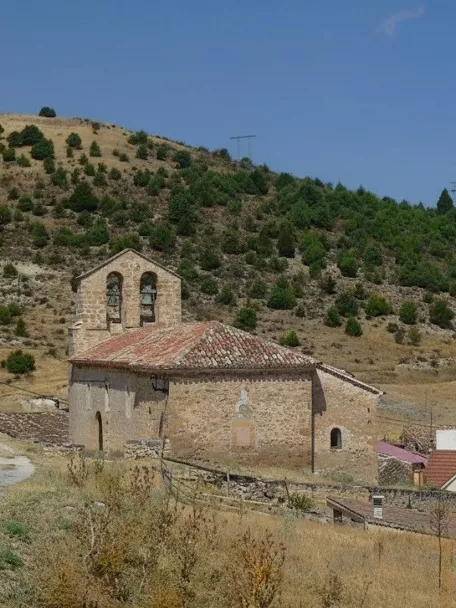 Pequeña iglesia de piedra en colina con casas al fondo y campanario de dos vanos.