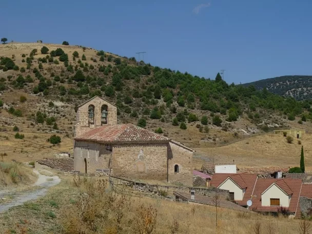 Pequeña iglesia de piedra en colina con casas al fondo y campanario de dos vanos.