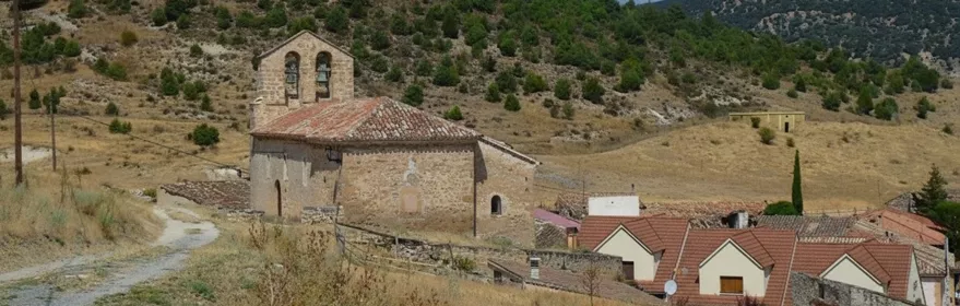 Pequeña iglesia de piedra en colina con casas al fondo y campanario de dos vanos.