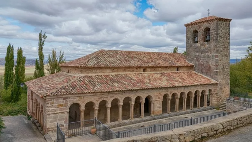 Iglesia románica de piedra con galería porticada y torre campanario bajo cielo nublado.