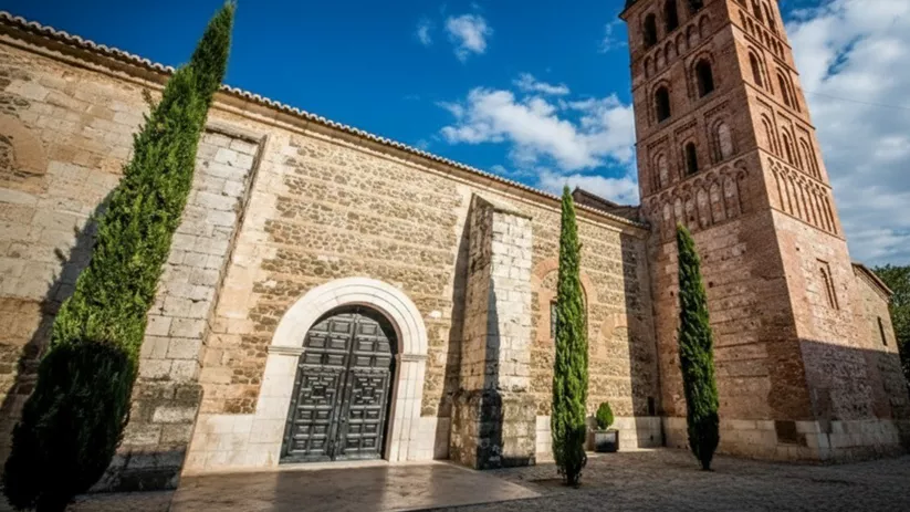 Fachada de iglesia de piedra y ladrillo con torre campanario y cipreses en la entrada.