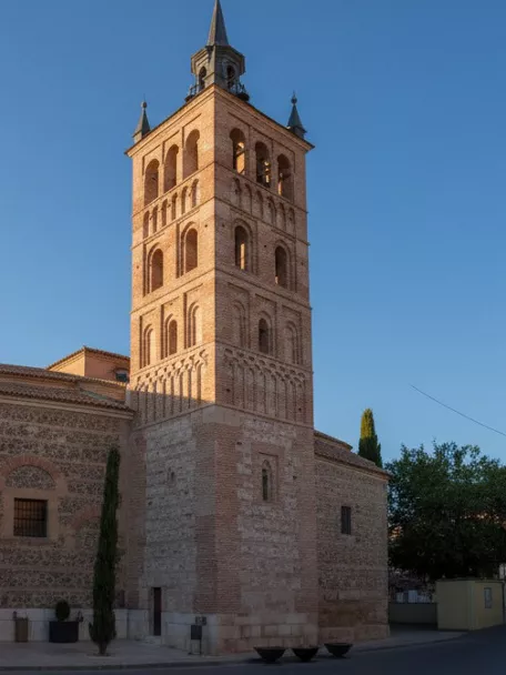 Vista lateral de iglesia con torre alta en esquina de una plaza urbana.