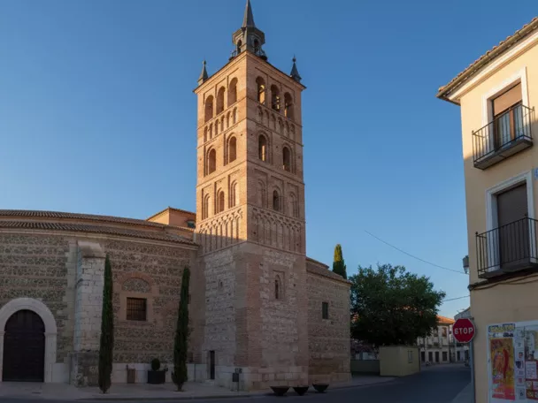 Vista lateral de iglesia con torre alta en esquina de una plaza urbana.