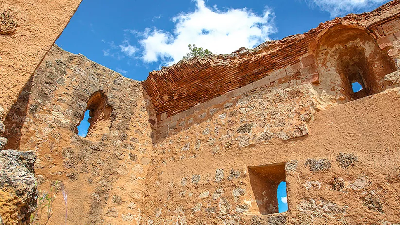 Interior de unas ruinas de piedra antiguas con muros erosionados, ventanas pequeñas y un arco parcial, abiertas al cielo azul con nubes blancas.