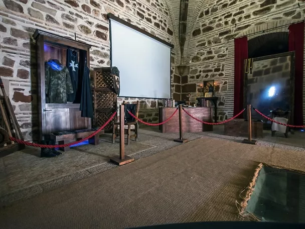 Interior de sala de piedra con exposición medieval y pantalla.