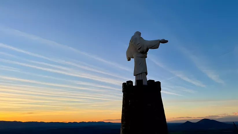 Estatua monumental con brazos abiertos sobre torre al atardecer