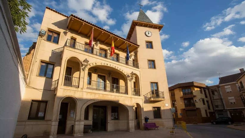 Casa Consistorial de Madrigueras con balcones, banderas oficiales y torre con reloj bajo un cielo parcialmente nublado.