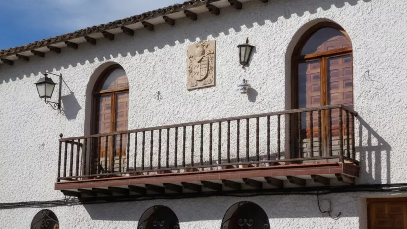 Fachada de una casa blanca con un balcón de madera, ventanas de arco de medio punto y un escudo heráldico en el centro.