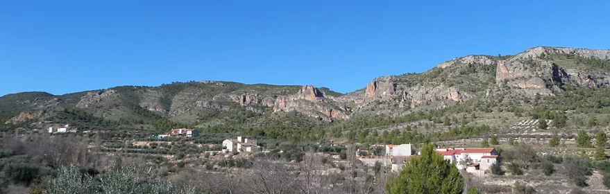 Valle rural con casas dispersas al pie de una sierra rocosa y cielo azul.