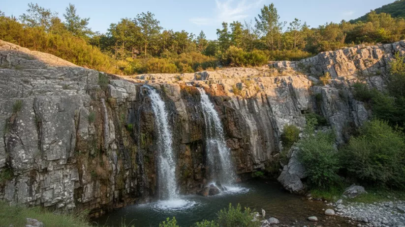 Doble cascada cayendo por una pared rocosa hacia una poza, con arbustos y piedras en la orilla.