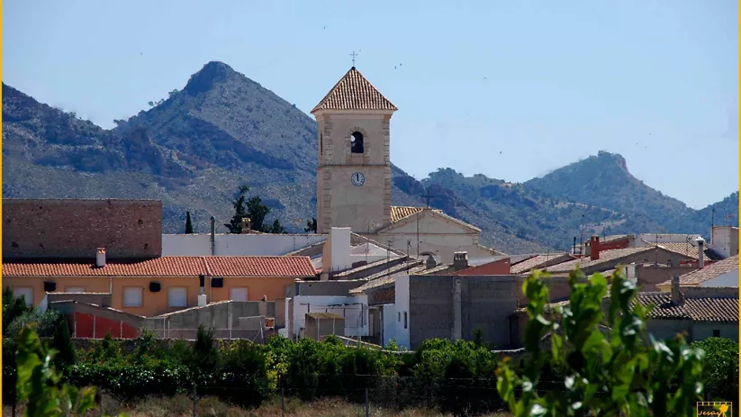 Iglesia con torre campanario entre viviendas, con montañas al fondo.