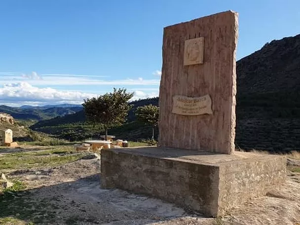 Monumento de piedra en un mirador con paisaje montañoso y pueblo a lo lejos.