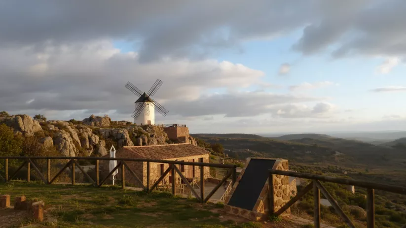 Mirador con barandilla y edificio de piedra frente a un molino de viento en la cima, con valle al fondo.