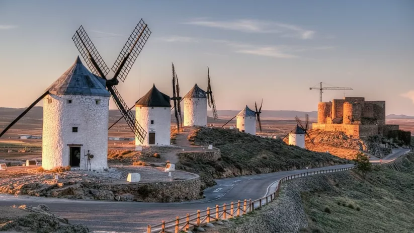 Molinos y castillo sobre colina con carretera y paisaje abierto al atardecer.