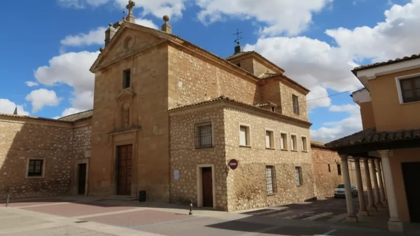 Iglesia de piedra en una plaza, con fachada rectangular y campanario bajo cielo con nubes.