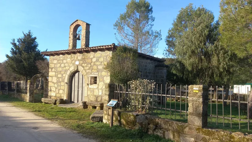Pequeña ermita de piedra con espadaña y verja entre árboles