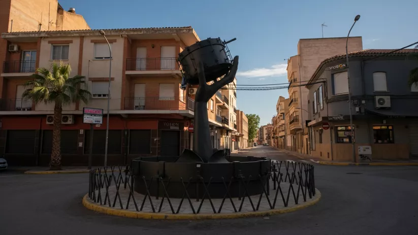 Rotonda con escultura metálica y tambor, en cruce urbano con comercios y viviendas.