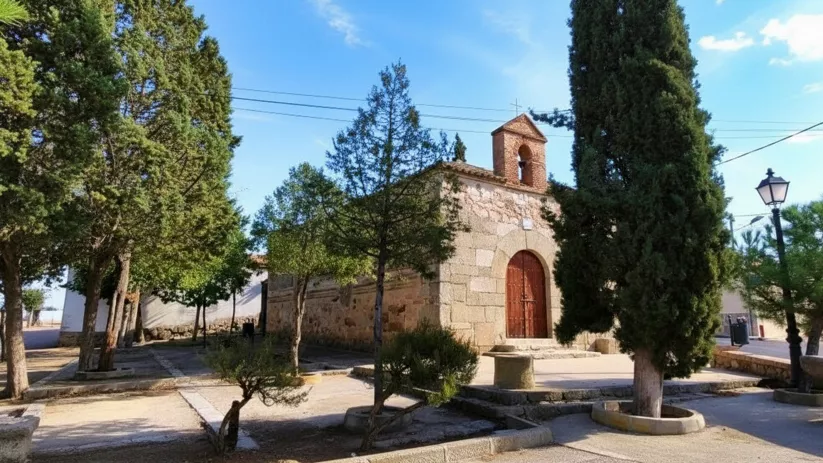 Pequeña iglesia de piedra en plaza arbolada con farola.