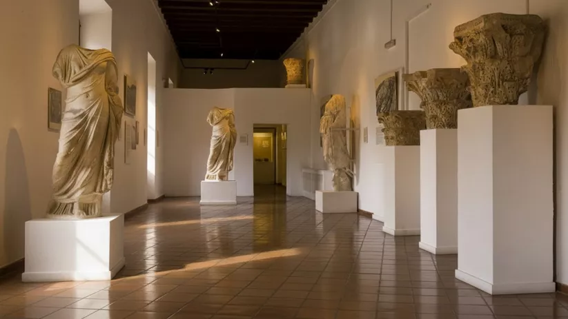 Sala con esculturas clásicas y capiteles sobre pedestales blancos.