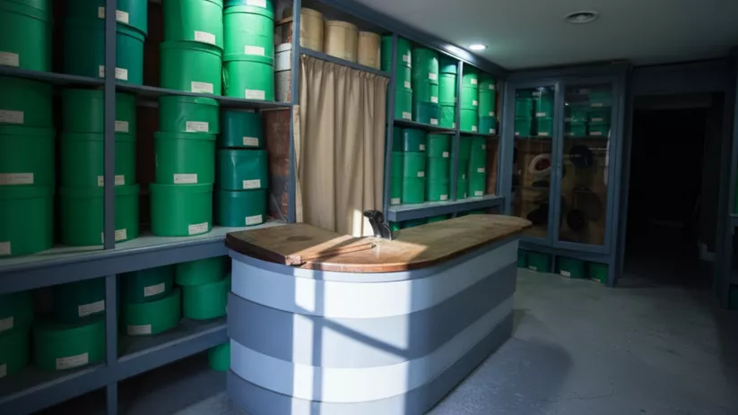 Almacén con estanterías llenas de recipientes verdes y un mostrador central.