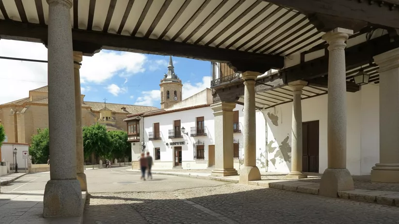 Vista desde un soportal hacia una plaza luminosa, con columnas de piedra y campanario al fondo.