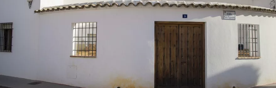 Fachada encalada en esquina con puerta de madera y ventanas con rejas.
