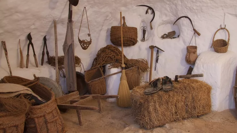 Sala con aperos tradicionales, cestos de esparto y herramientas colgadas en pared blanca.