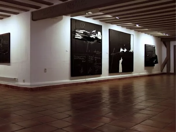 Sala amplia con pinturas oscuras sobre paredes blancas y suelo de barro.
