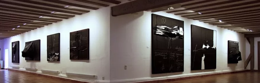 Sala amplia con pinturas oscuras sobre paredes blancas y suelo de barro.