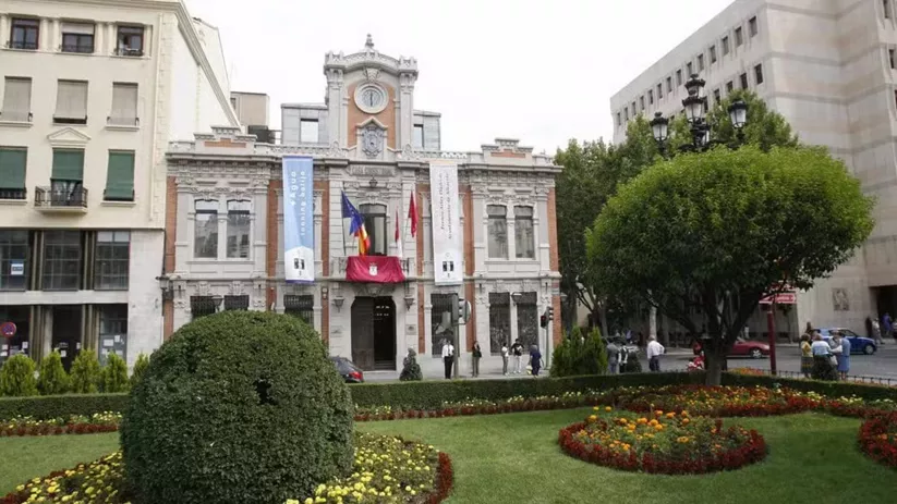 Edificio histórico con fachada ornamental y jardín delantero con flores y setos.