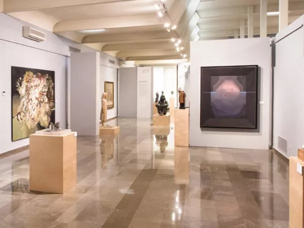 Sala de exposiciones con cuadros y esculturas distribuidos en un espacio amplio y luminoso.