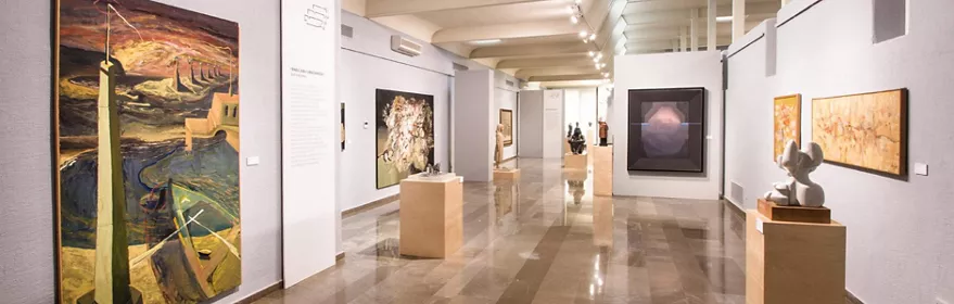 Sala de exposiciones con cuadros y esculturas distribuidos en un espacio amplio y luminoso.