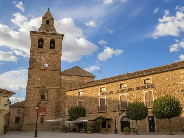 Plaza con torre de piedra y edificio histórico bajo cielo azul con nubes.